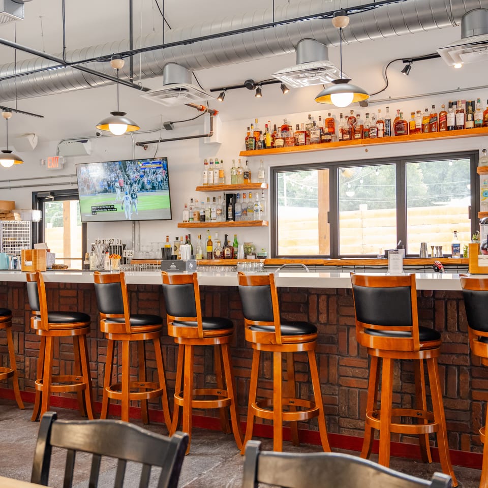 Restaurant bar with wooden stools, exposed ductwork, liquor shelves, large windows, and a television showing a soccer game.
