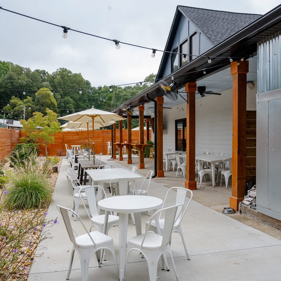 Outdoor restaurant patio with white tables, chairs, umbrellas, string lights, wooden posts, garden landscaping, and wooded surroundings.