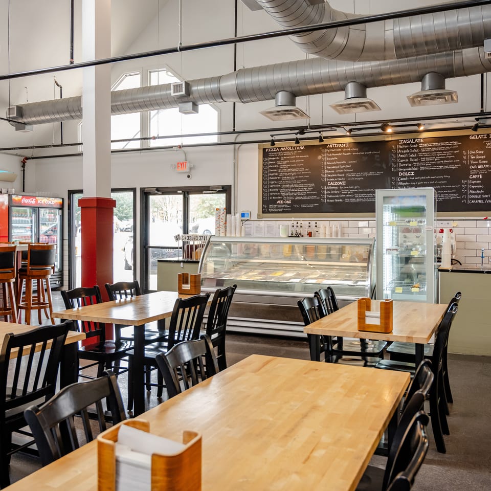 Bright Italian restaurant interior with wooden tables, black chairs, gelato display case, chalkboard menu, and exposed ventilation ducts.