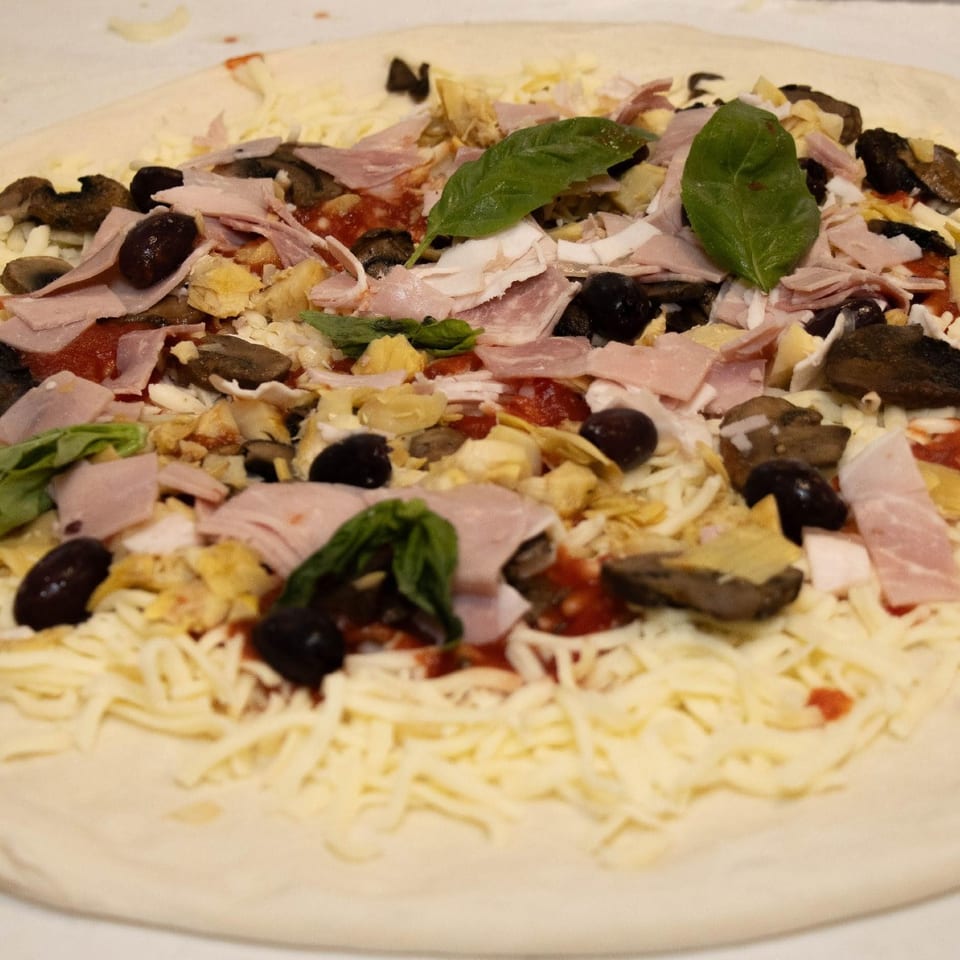 Pizza topped with ham, mushrooms, olives, artichokes, and basil before baking.