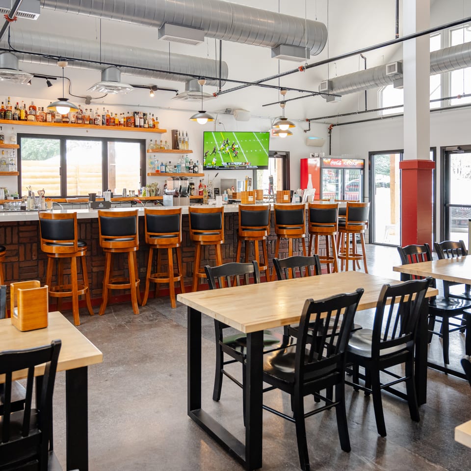 Bright modern restaurant bar with liquor shelves, wooden tables, black chairs, pendant lights, exposed ductwork, and a television showing football.