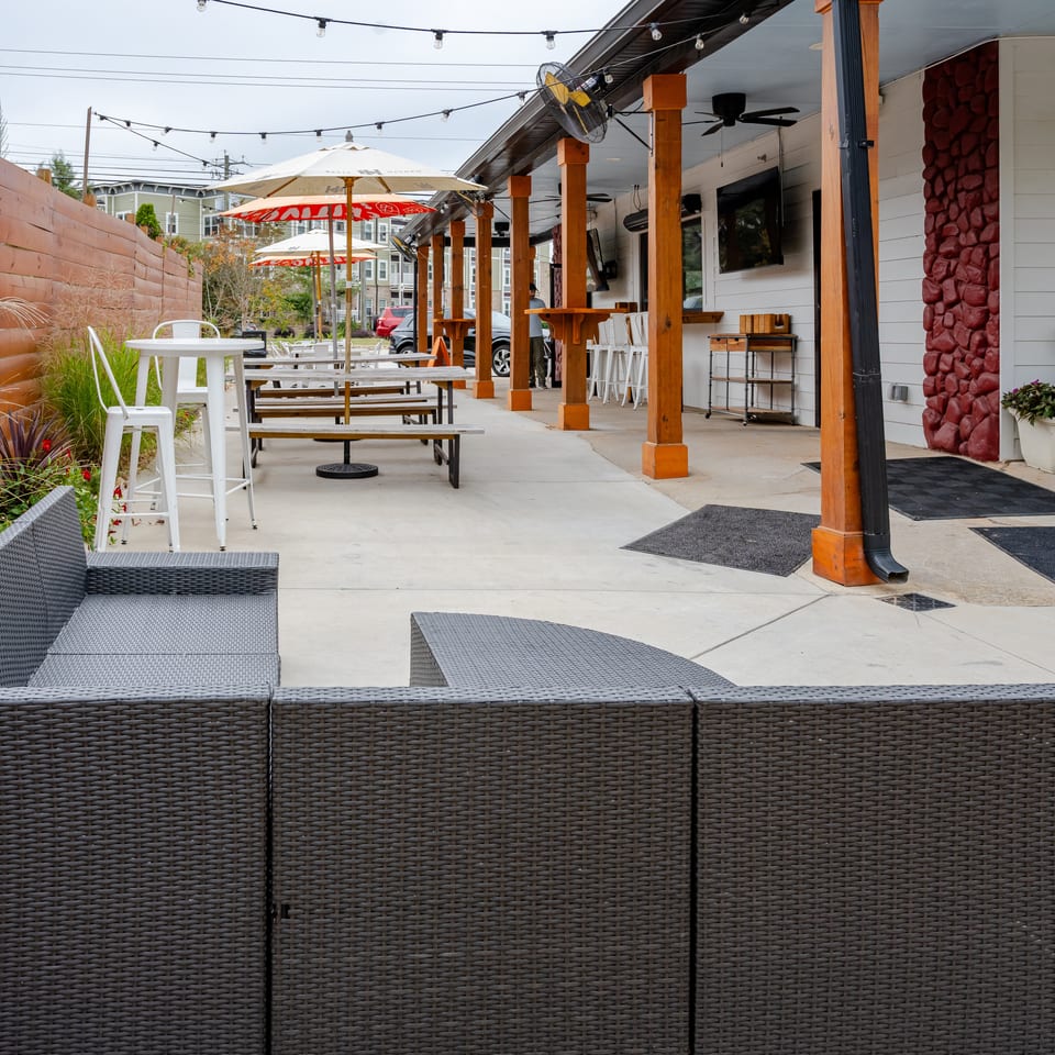 Outdoor restaurant patio with picnic tables, umbrellas, string lights, wicker seating, and landscaped planters beside a covered dining area.