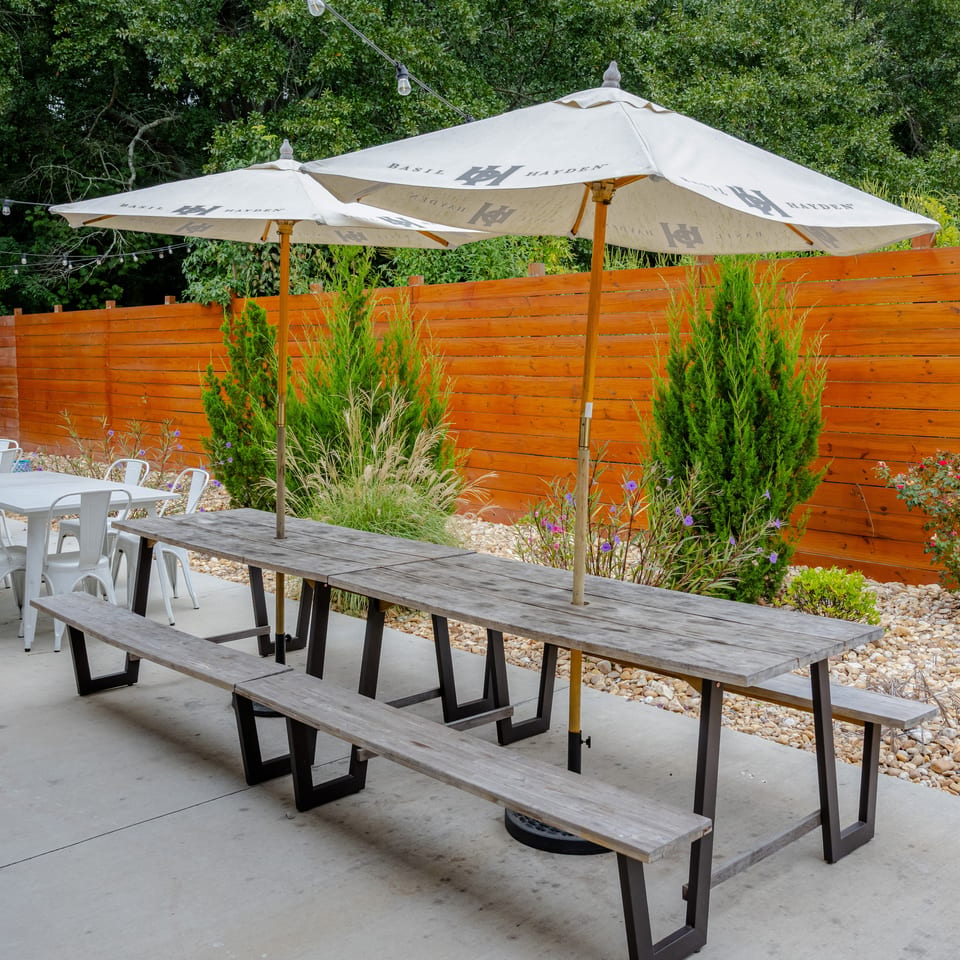 Outdoor restaurant patio with picnic tables, white chairs, branded umbrellas, string lights, greenery, and a wooden privacy fence.