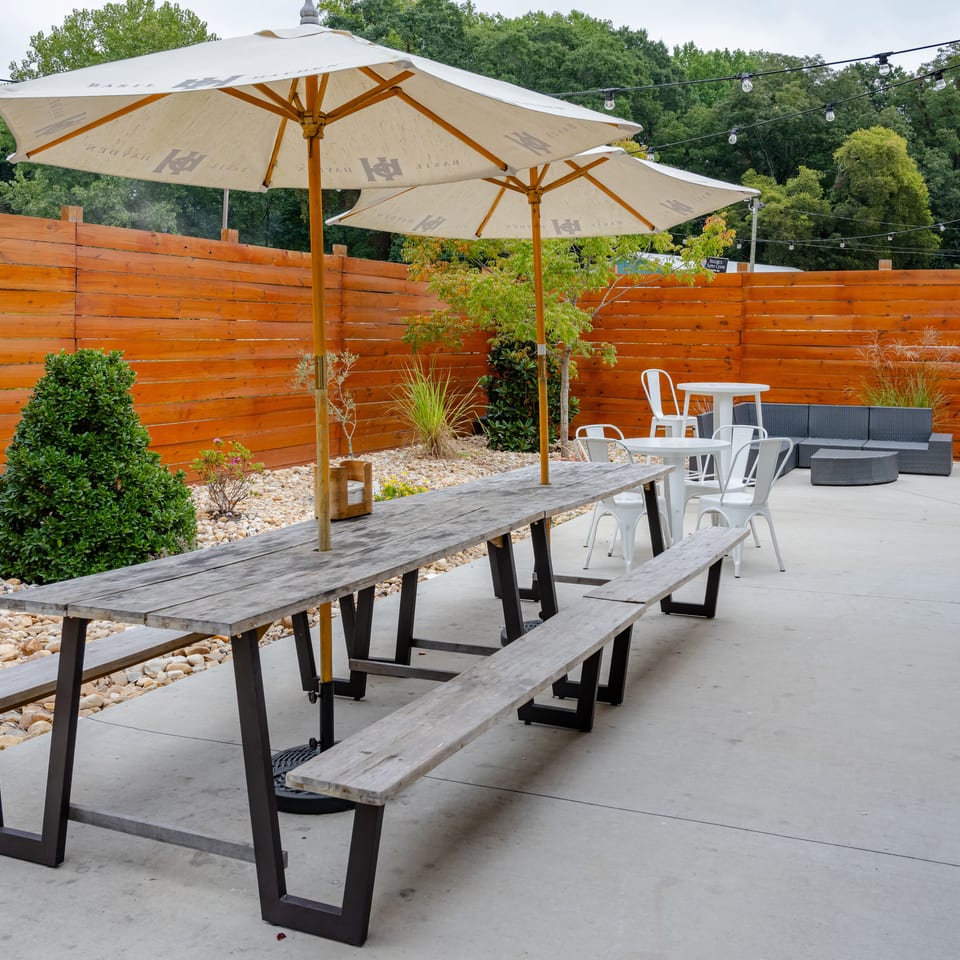 Outdoor restaurant patio with picnic tables, white umbrellas, café seating, string lights, landscaping, and wooden privacy fencing.