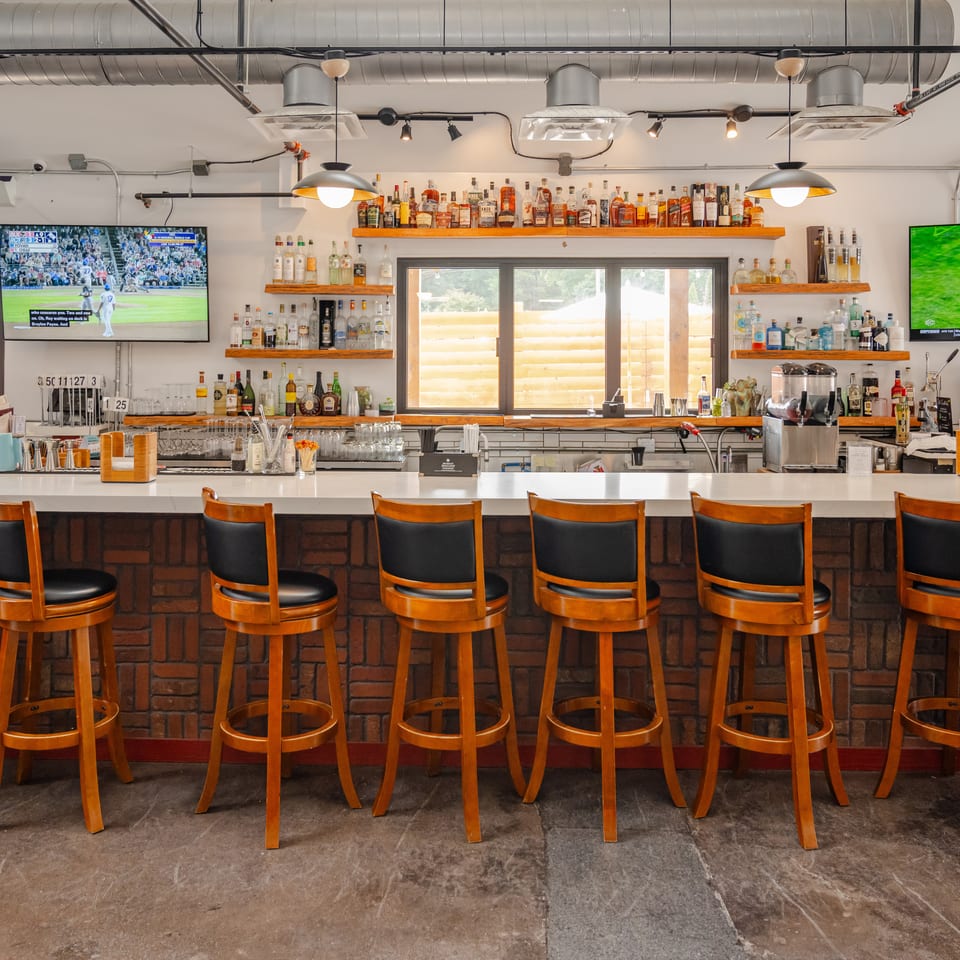 Spacious restaurant bar with wooden stools, liquor shelves, pendant lighting, exposed ductwork, and televisions showing sports.