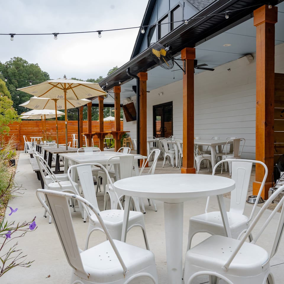 Outdoor restaurant patio with white tables, metal chairs, umbrellas, string lights, wooden posts, and landscaped greenery.