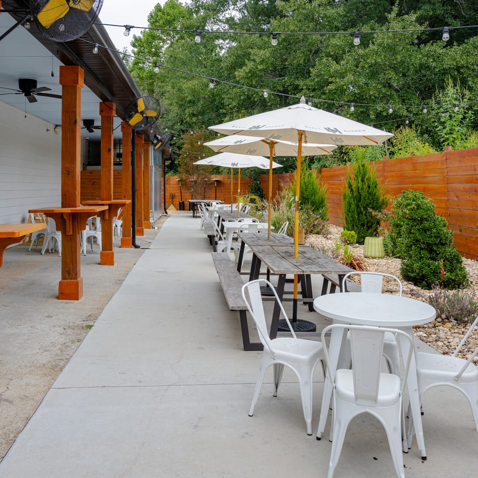 Outdoor restaurant patio with picnic tables, white chairs, umbrellas, string lights, fans, and landscaped garden beside a wooden fence.
