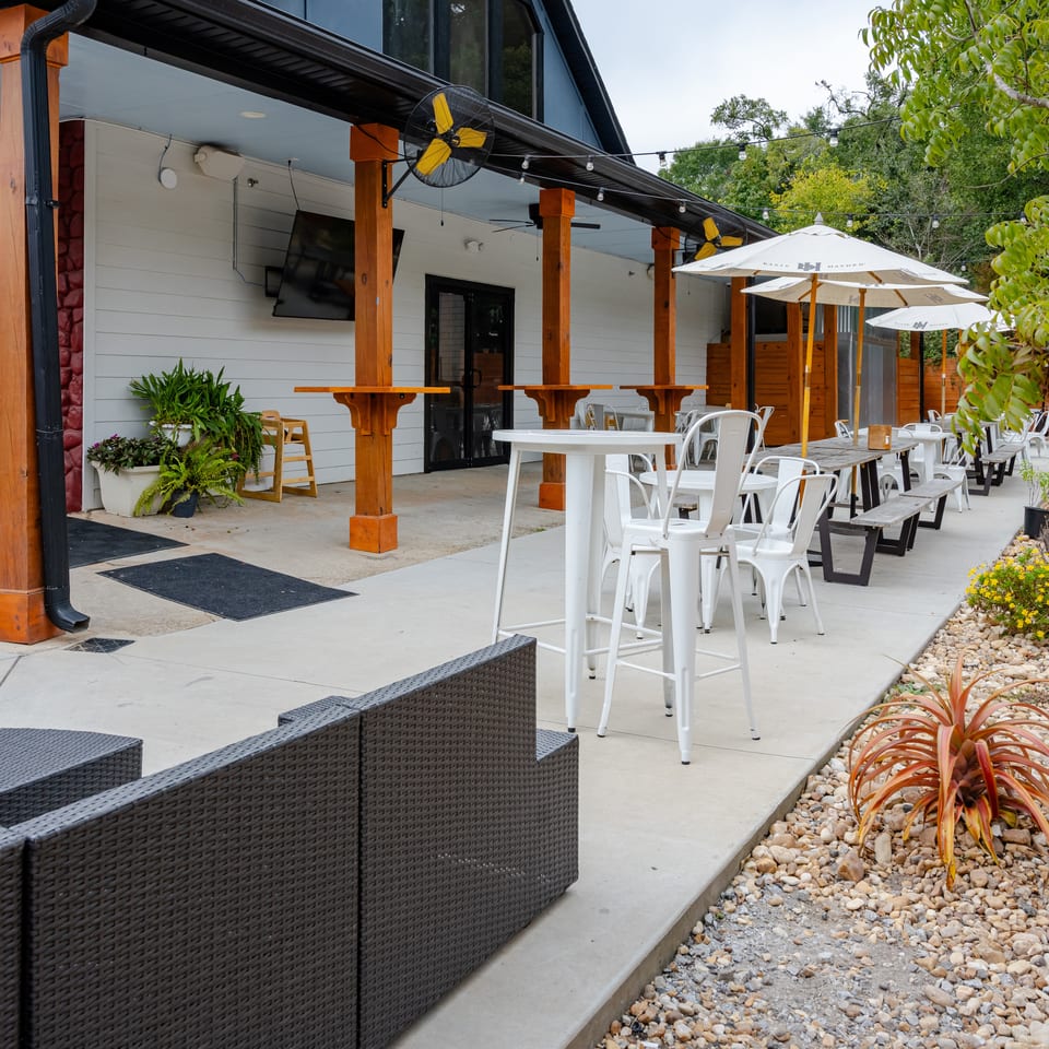 Outdoor restaurant patio with wooden posts, white umbrellas, tables, chairs, fans, string lights, and landscaped rock garden.