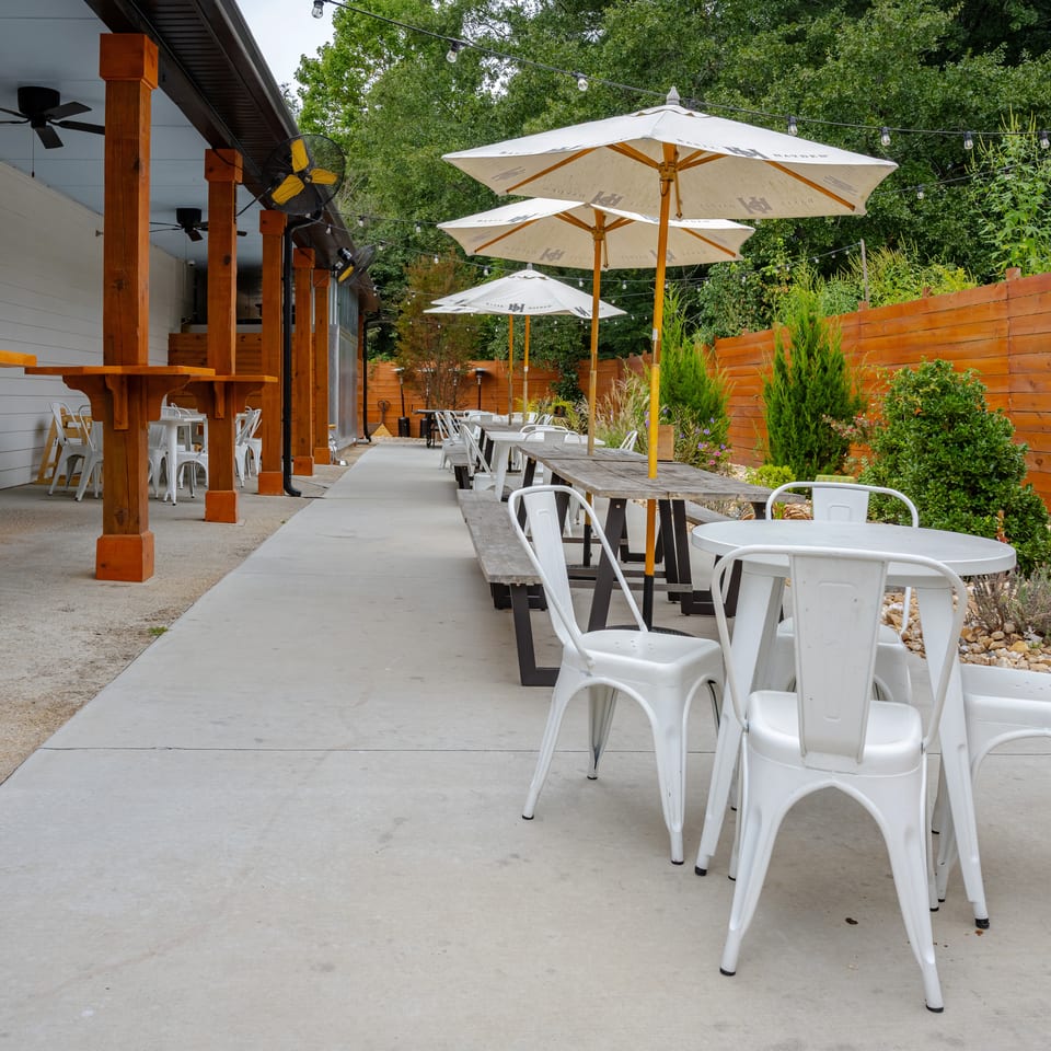 Outdoor restaurant patio with white tables and chairs, large umbrellas, string lights, wooden accents, and lush greenery.