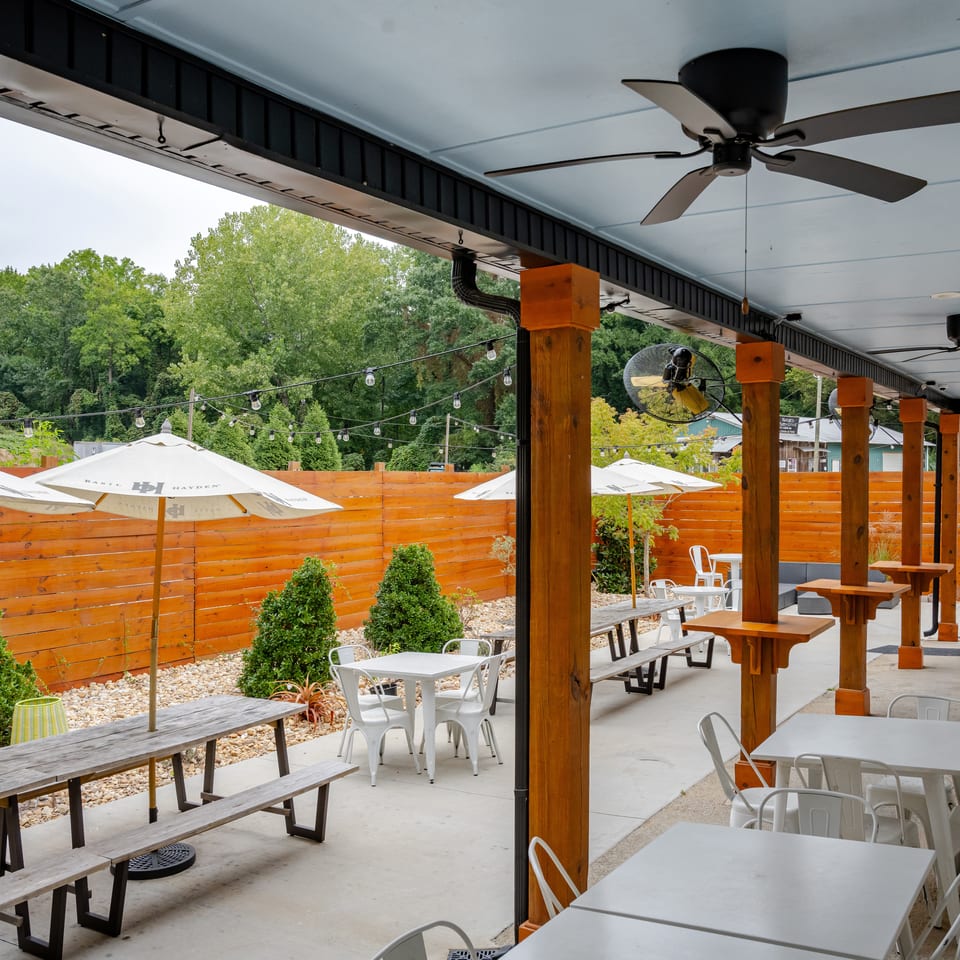 Covered restaurant patio with wooden beams, ceiling fans, string lights, picnic tables, umbrellas, and surrounding trees.