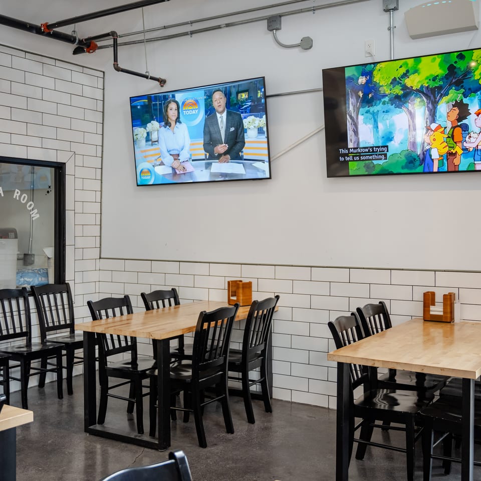 Bright restaurant dining area with subway-tile walls, wooden tables, black chairs, and two televisions displaying news and Pokémon.