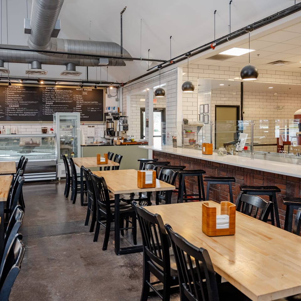 Spacious Italian restaurant dining room with wooden tables, black chairs, tiled service counter, gelato case, and chalkboard menu.