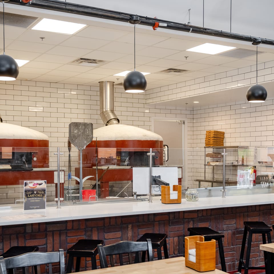 Restaurant interior with brick counter, tiled walls, wood-fired ovens, pendant lights, and dining tables.