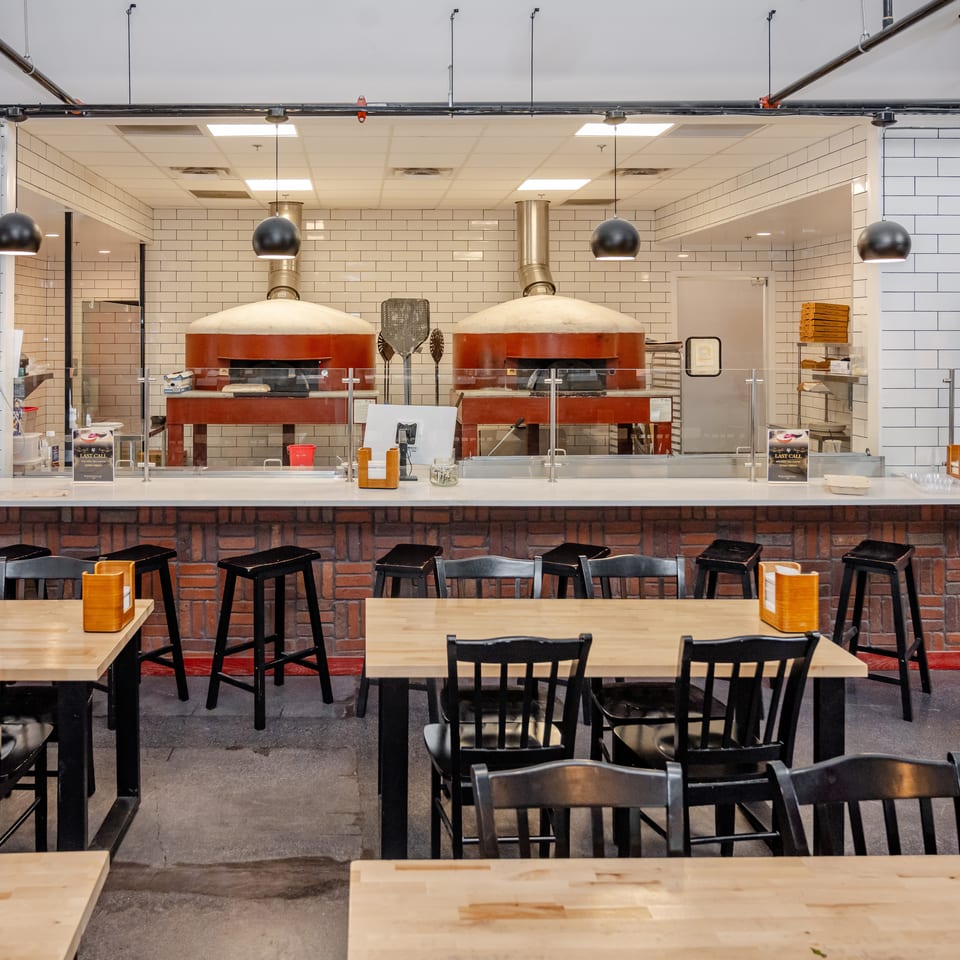 Spacious restaurant dining area with wooden tables, black chairs, tiled walls, and two large red-brick pizza ovens behind the counter.