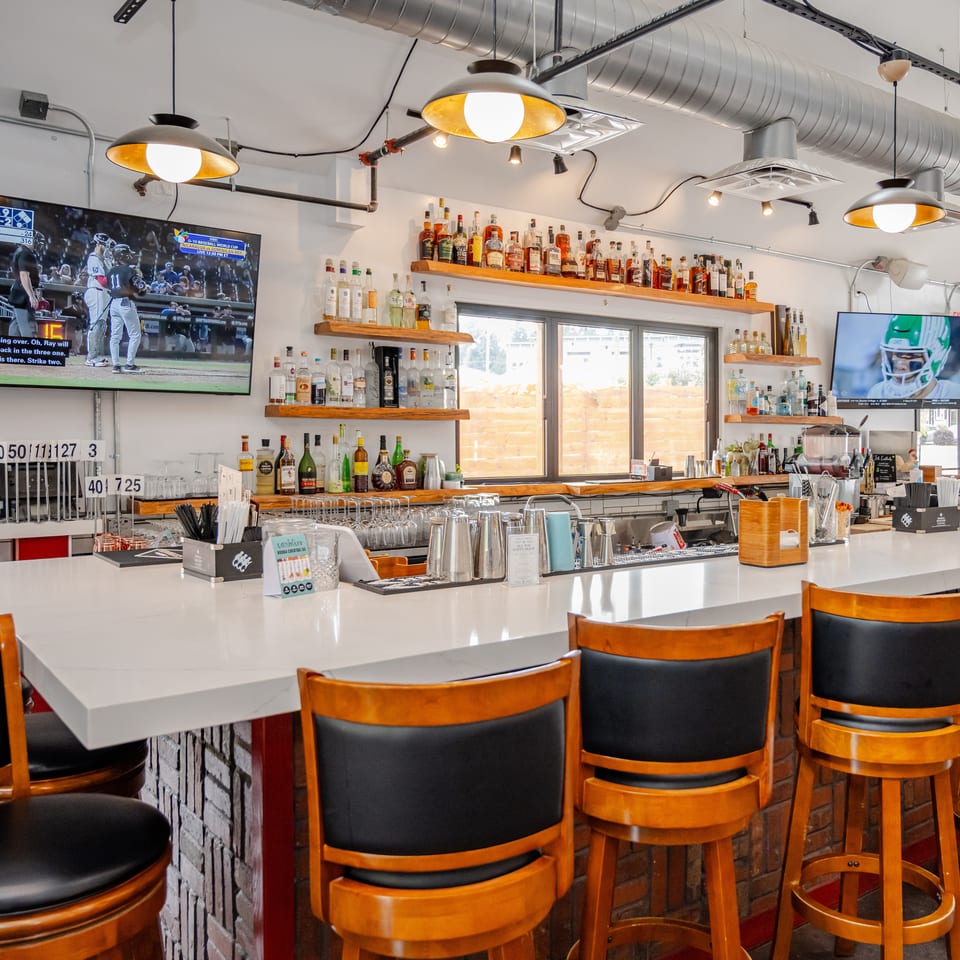 Bright restaurant bar with liquor shelves, high-top seating, exposed ductwork, and televisions showing sports.