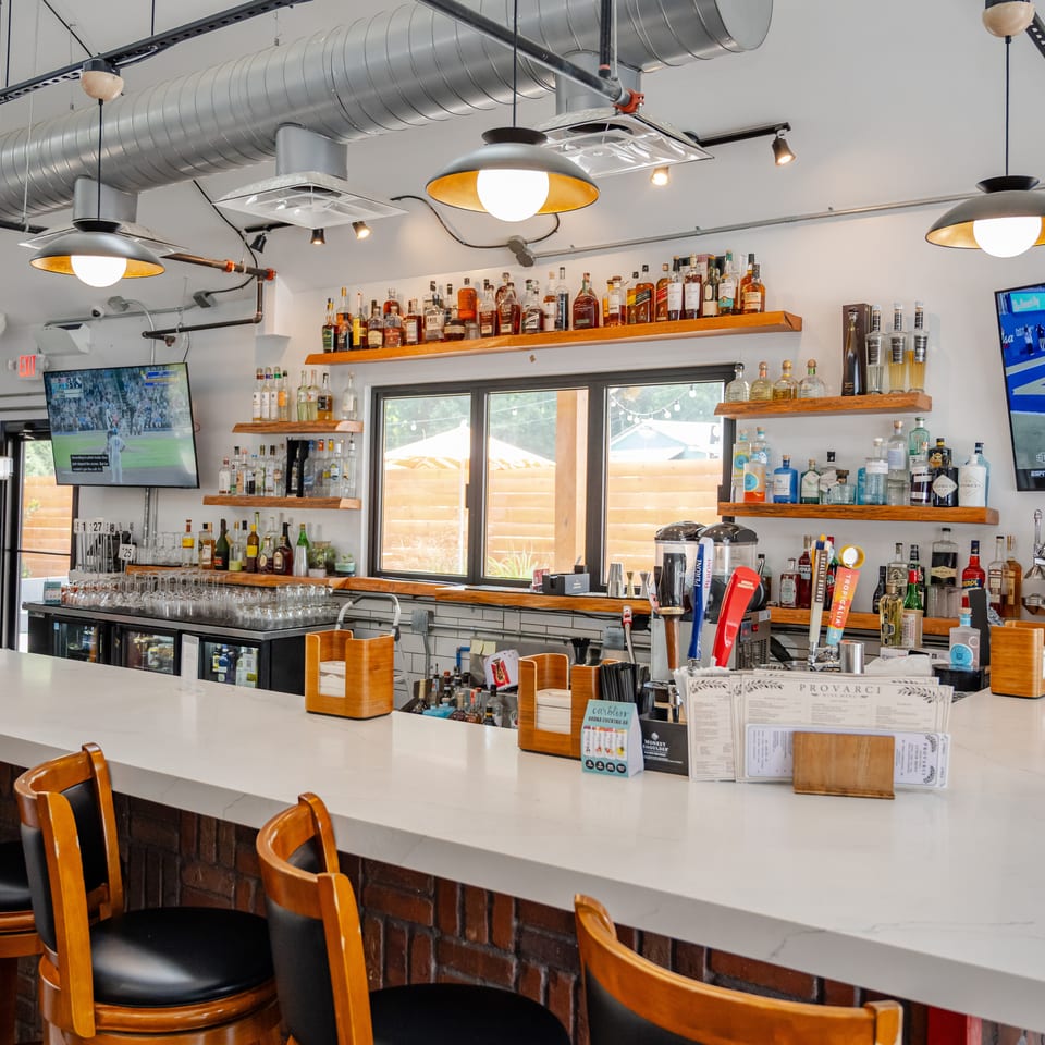 Bright restaurant bar with wooden liquor shelves, white countertop, barstools, pendant lights, and televisions showing sports.